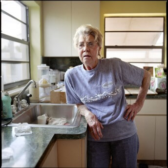 A still from The Graying of AIDS. A person with short blonde hair, jeans, and a purple t-shirt stands in a Kitchen and leans over the sink.