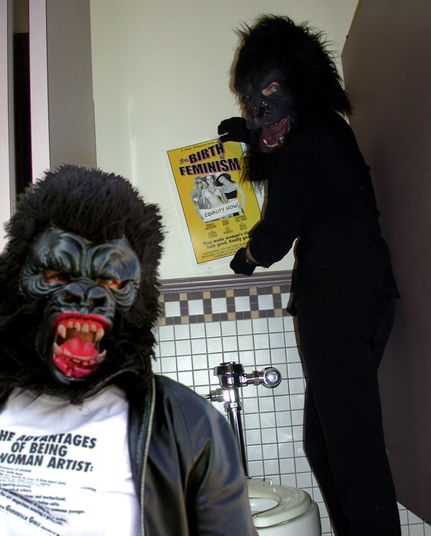 Two people in gorilla suits and face masks. One is sitting and the other stands behind them touching a poster on the wall.