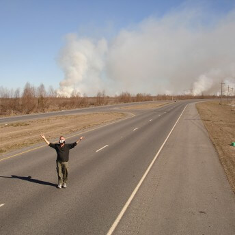 Still from My Louisiana Love. A man, wearing an air filtering mask, stands in the middle of a 4 lane road. There are multiple clouds of smoke in the background.