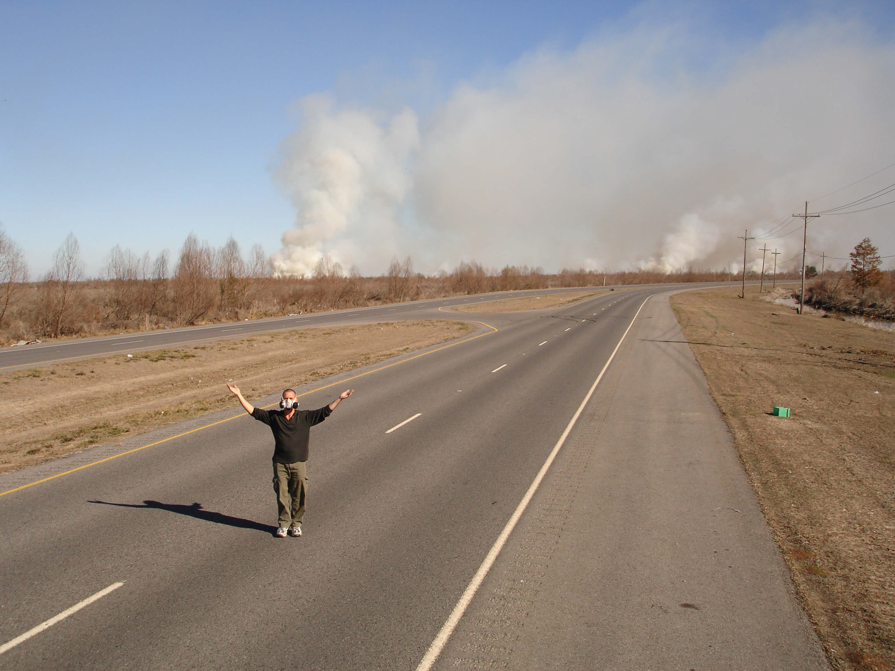 Still from My Louisiana Love. A man, wearing an air filtering mask, stands in the middle of a 4 lane road. There are multiple clouds of smoke in the background.