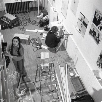 An aerial shot of two people in a room with a ladder, wires, and electronics on the floor. Photographs are on the wall. Black and white photograph.