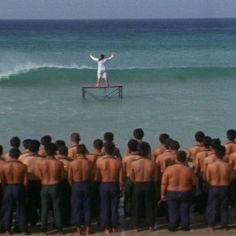 Still from Our City Dreams. There is a woman in a white dress standing on a platform in an ocean's surf with her hands raised in the air. On the waters edge, many shirtless men stand in rows looking towards the ocean.