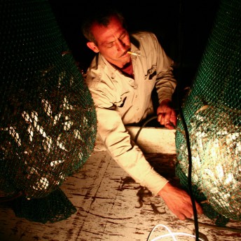 Still from My Louisiana Love. A man smoking a cigarette untangles a wire. There are two large nets filled with fish.