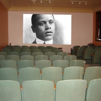 Still from Oscar's Comeback. A theatre space with green metal chairs facing the wall with the projection of a portrait of a man in black and white.