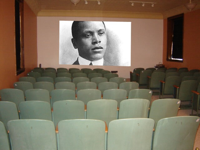 Still from Oscar's Comeback. A theatre space with green metal chairs facing the wall with the projection of a portrait of a man in black and white.