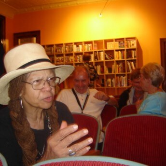 Still from Oscar's Comeback. A woman in a hat is speaking to someone off camera. She is seated in a red velvet theatre chair surrounded by others. There is a large bookshelf in the background as well.