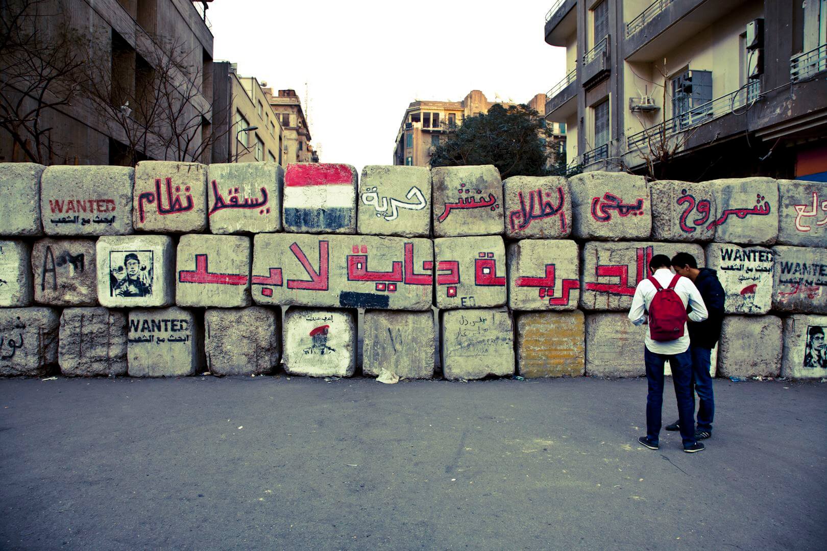 Large stacked blocks forming a wall covered in spray painted Arabic text. Two people are standing to the right. Residential buildings stand behind the wall.