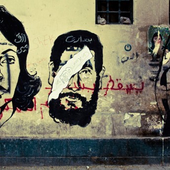 Two faces painted with a black outline and red Arabic writing on a yellow wall. A cutoff caged window lies above the street art.