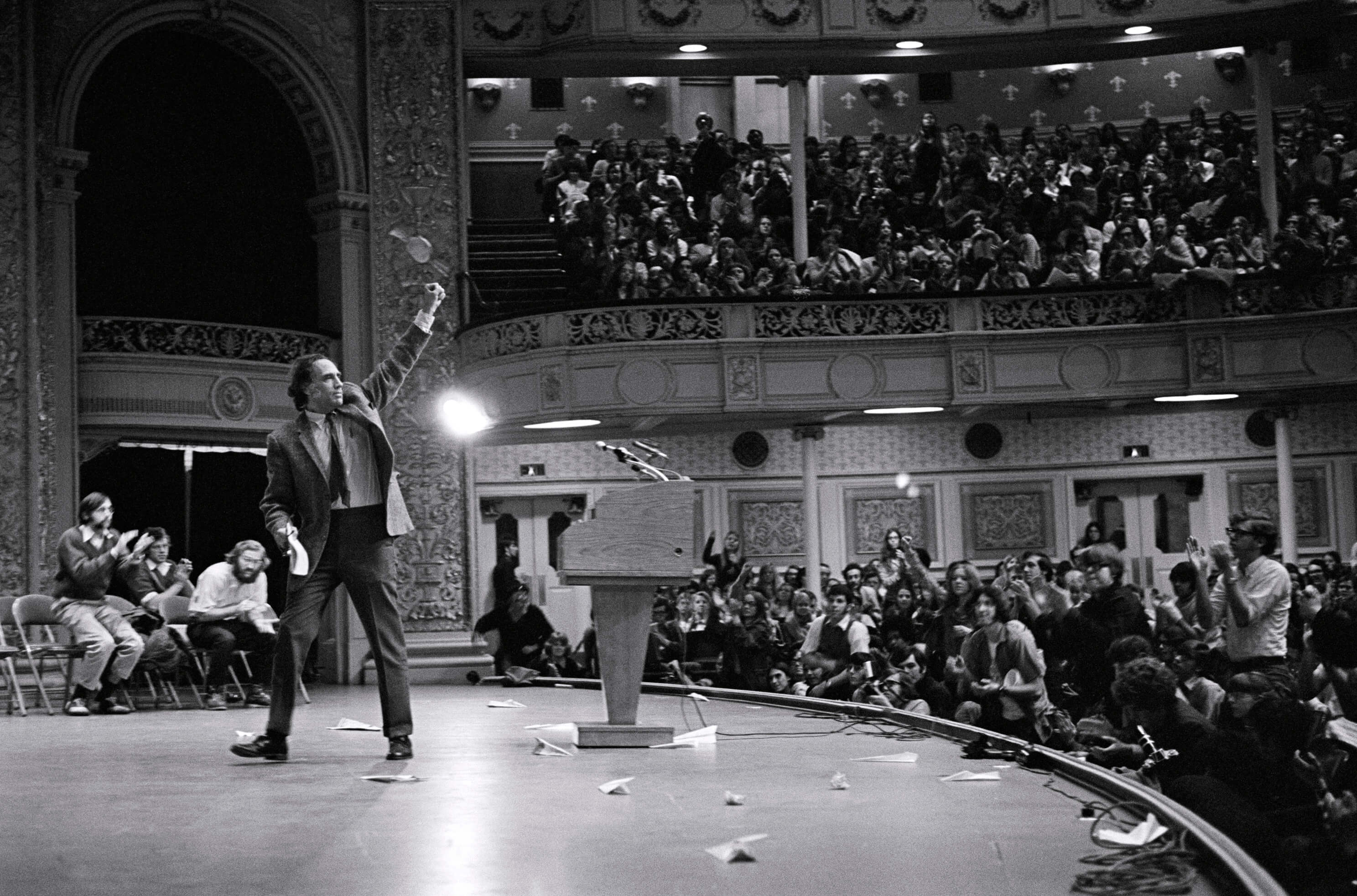 William Kunstler raises a fist to the sky on a stage in front of a podium. The crowd of people in the background also stands. Black and white photograph.