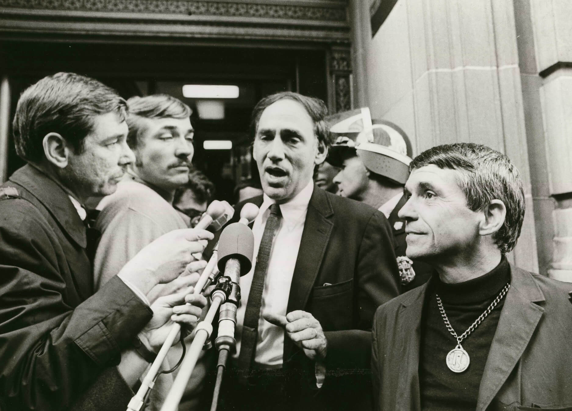 William Kunstler speaking into many microphones, surrounded by several people. Black and white photograph.