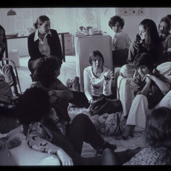 A group of people sits together. A few sit on rugs on the floor, while others sit in chairs and on the sofa.