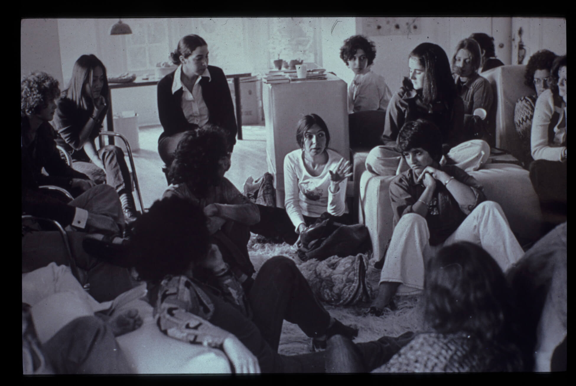 A group of people sits together. A few sit on rugs on the floor, while others sit in chairs and on the sofa.