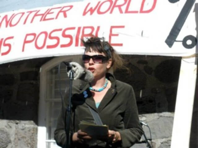 Still from The Mouse That Roared. A person wearing sunglasses stands outside in front of a microphone. They are holding a small stack of papers in their hands. Behind them, a large banner reads: "-nother World Possible".