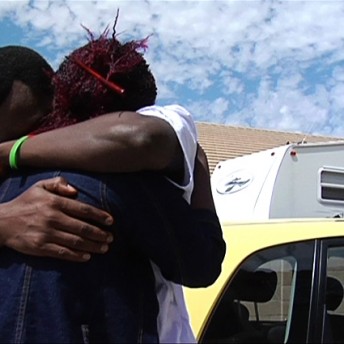 Still from Pushing The Elephant. Two people embrace outside next to vehicles.