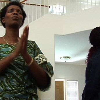 Still from Pushing The Elephant. Two women stand in a house. One of them has their eyes closed and her hands touching each other. The other one looks away from the camera.