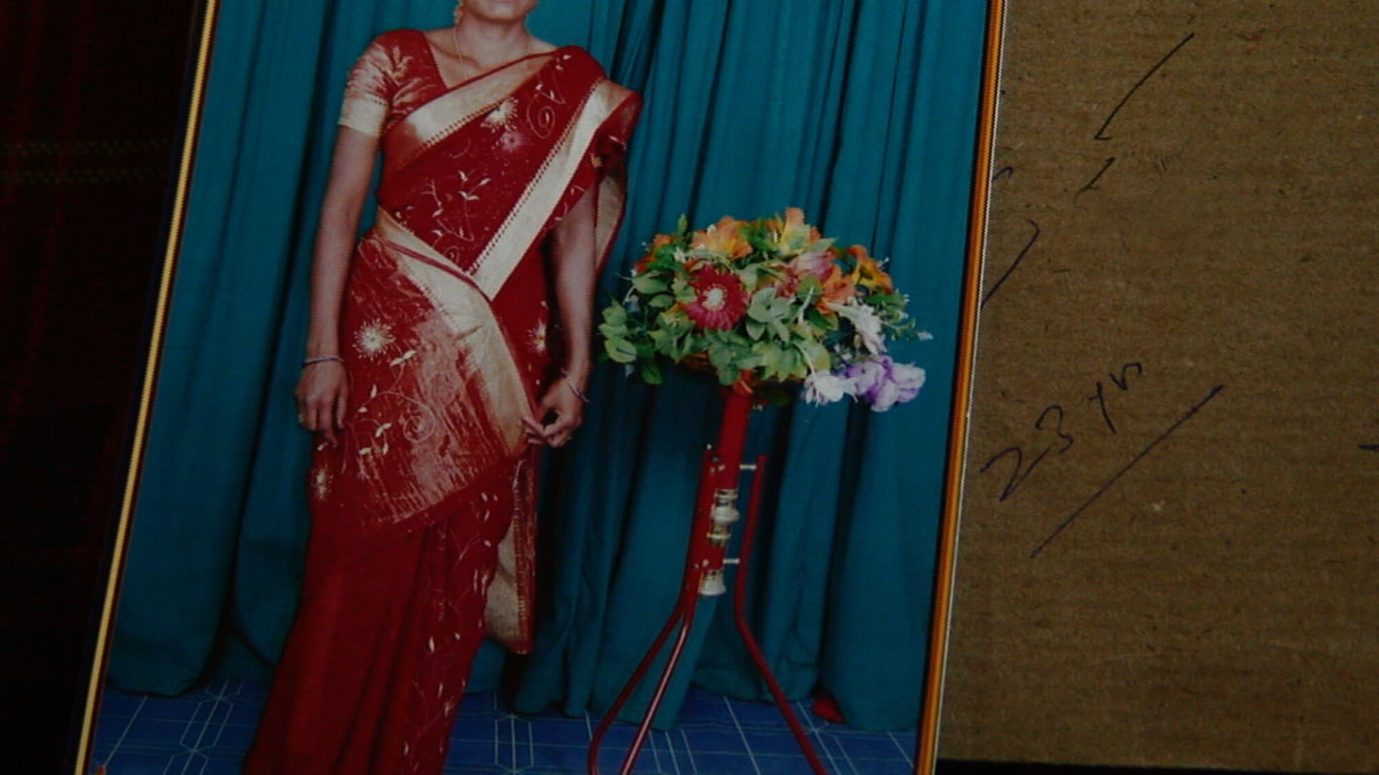 A framed portrait of a woman in a red sari standing beside a bouquet of flowers placed on a cardboard surface with handwriting that says "23 yr" to the right of the frame.