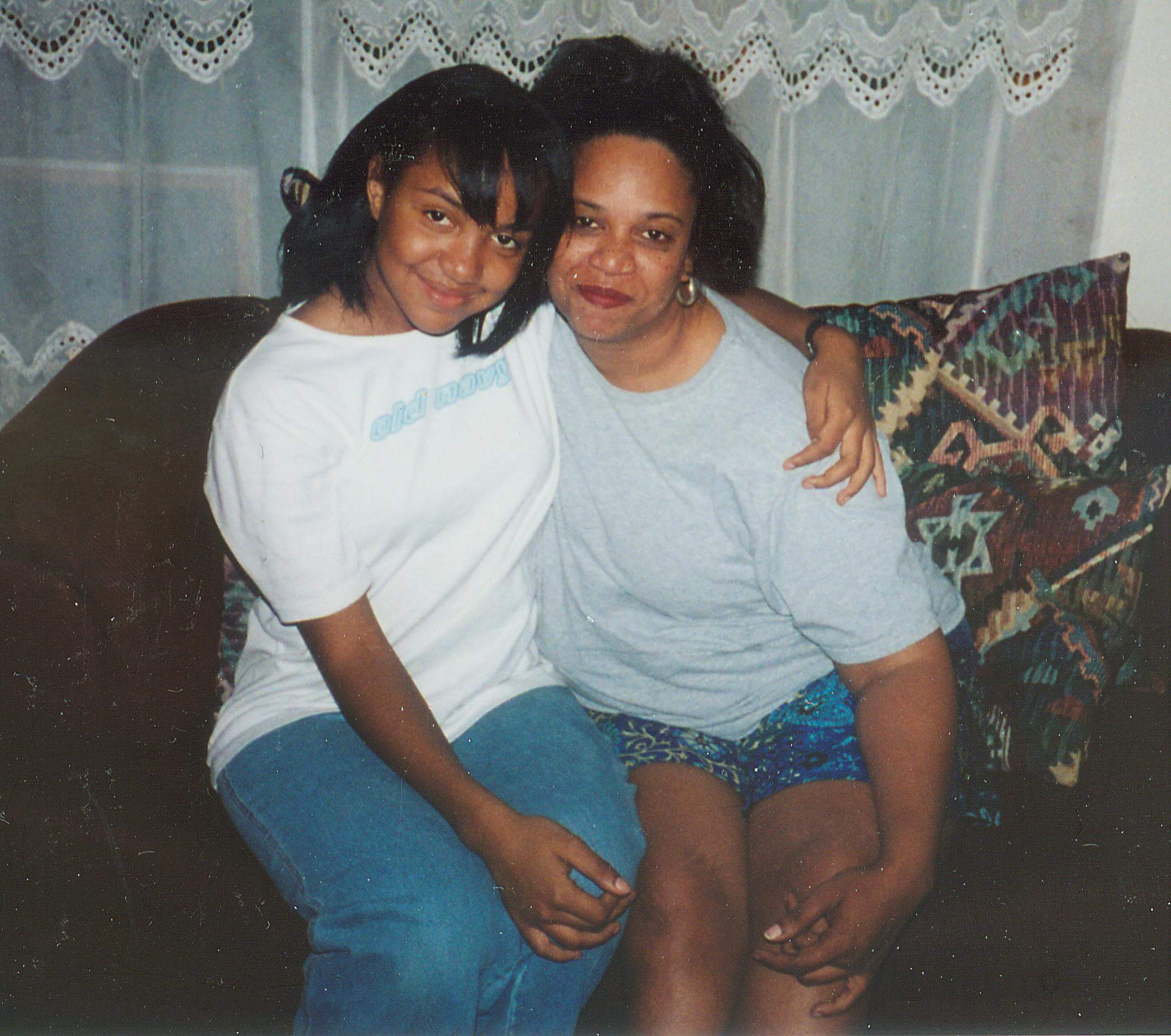 A teenager and a woman sitting on a sofa hug each other while posing in front of the camera.