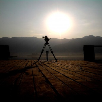 Still for I Never Left. A tripod, with its leg extended, is framed at the center of the image. A bright sun is in the sky over the mountains in the distance. The tripod casts a long shadow on a wooden deck.