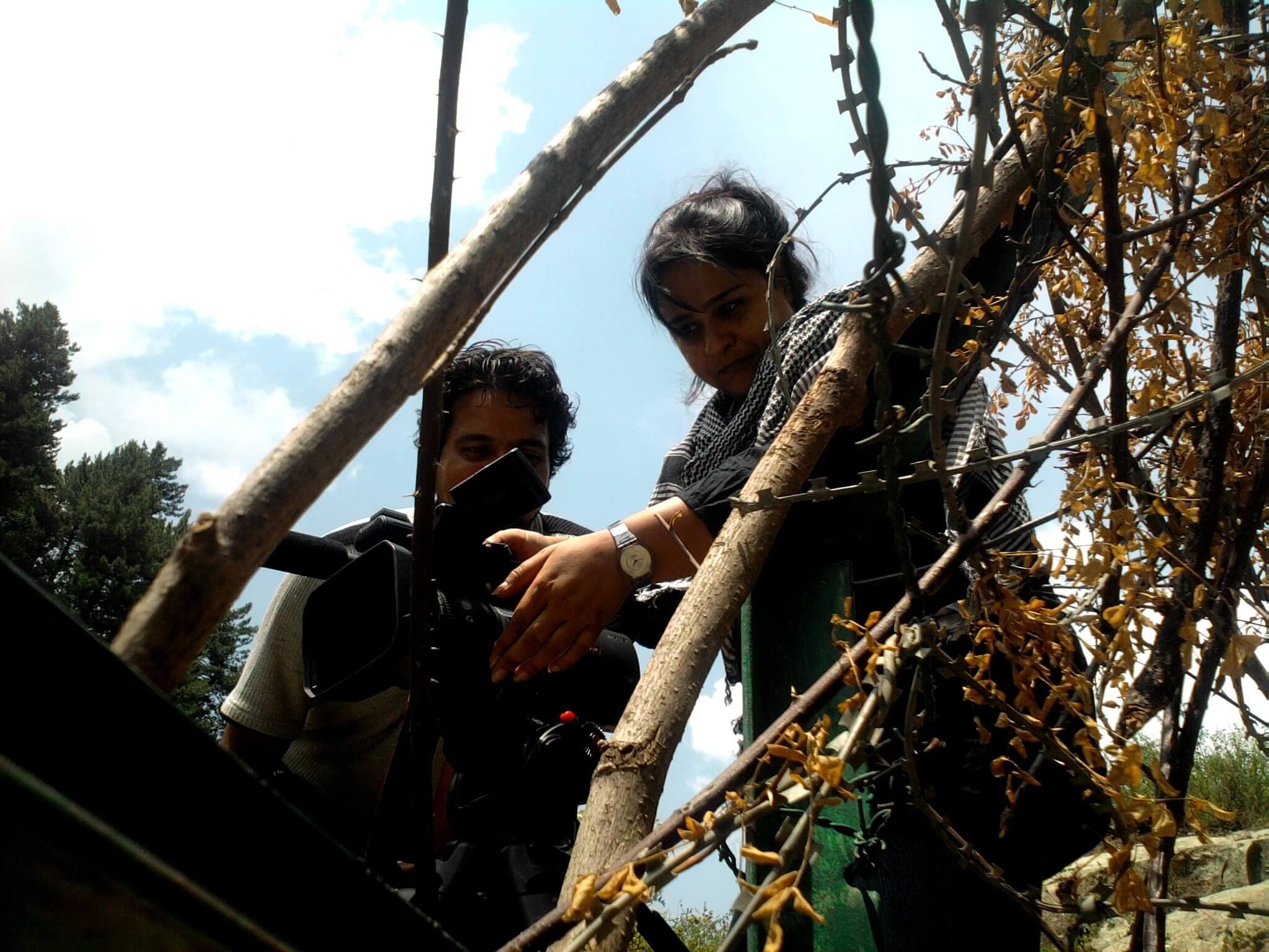 Production still from I Never Left. A woman positions a camera on a tripod. She is seen from a low angle in between branches. A man is looking on.