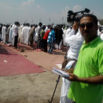 Still from I Never Left. A man in dark glasses and a bright green shirt is looking directly at the camera. Behind him, a person is filming with a large camera on his shoulder a group of people standing in lines in front of prayer mats.