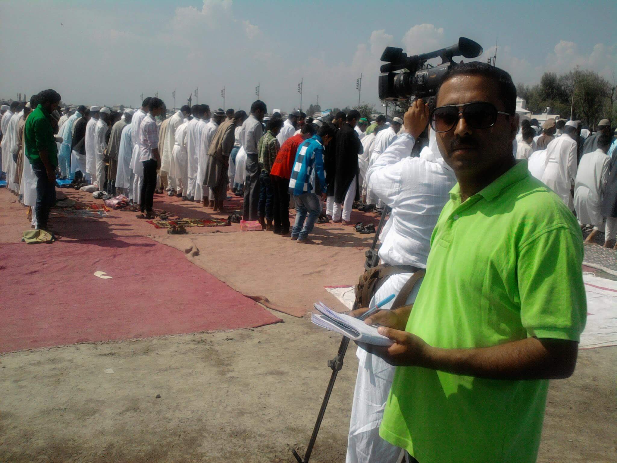 Still from I Never Left. A man in dark glasses and a bright green shirt is looking directly at the camera. Behind him, a person is filming with a large camera on his shoulder a group of people standing in lines in front of prayer mats.
