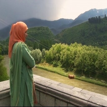 Still from I Never Left. A woman in a green salwar kameez and an orange hijab is standing with arms crossed on a stone balcony. She is looking down at a green field below her. There is a grove of trees at the edge of the field, and dark mountains and an overcast sky are seen in the distance.