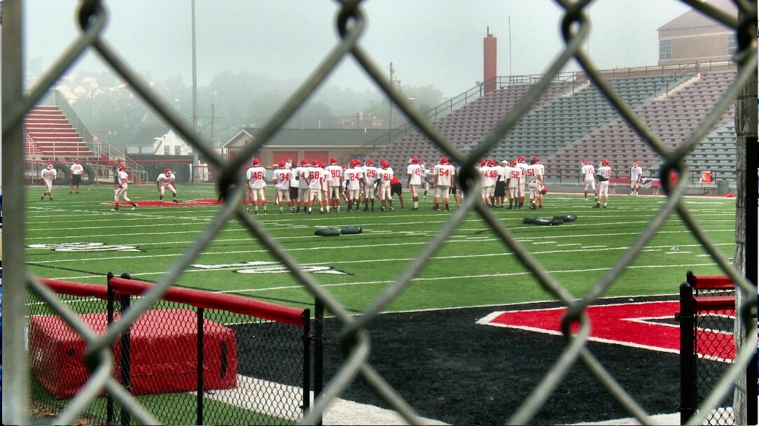 Still from Roll Red Roll. The shot was taken through a steel fence in the foreground, in the background a football team with white jerseys and red helmets practices on a football field.