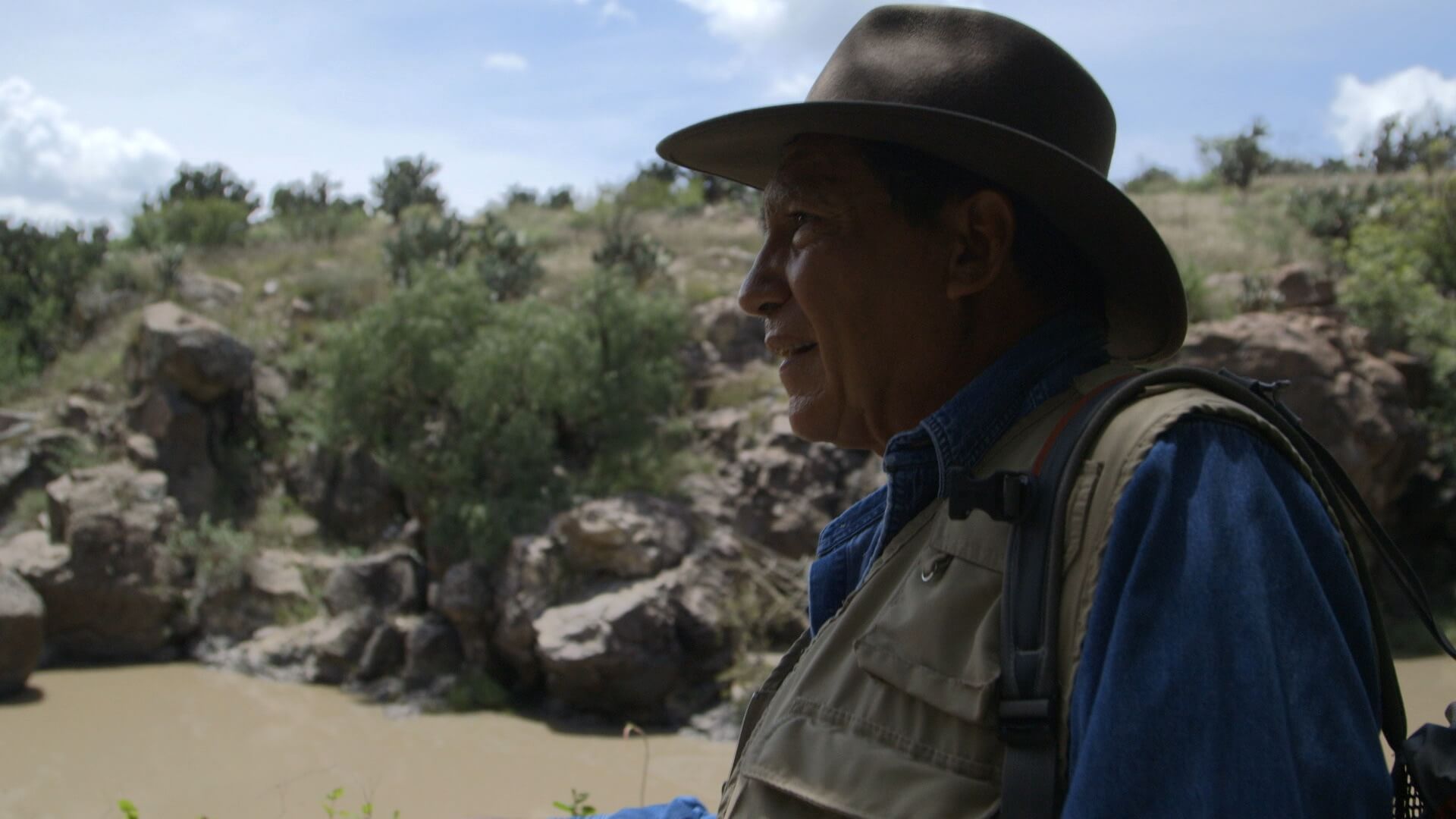 A still from The Age of Water. A man wears a large-brimmed hat and a vest, he stares off in the distance. He is standing in front of a river bank.