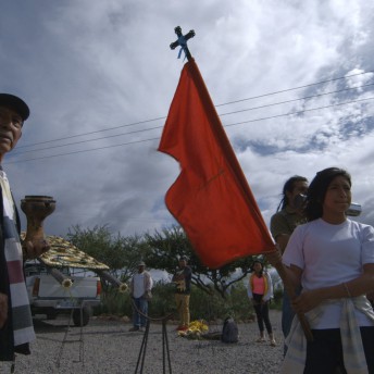 A still from The Age of Water. A person in a white T-shirt is holding a tall red flag with a cross atop the mast. Four other people stand behind them, in a patch of gravel.