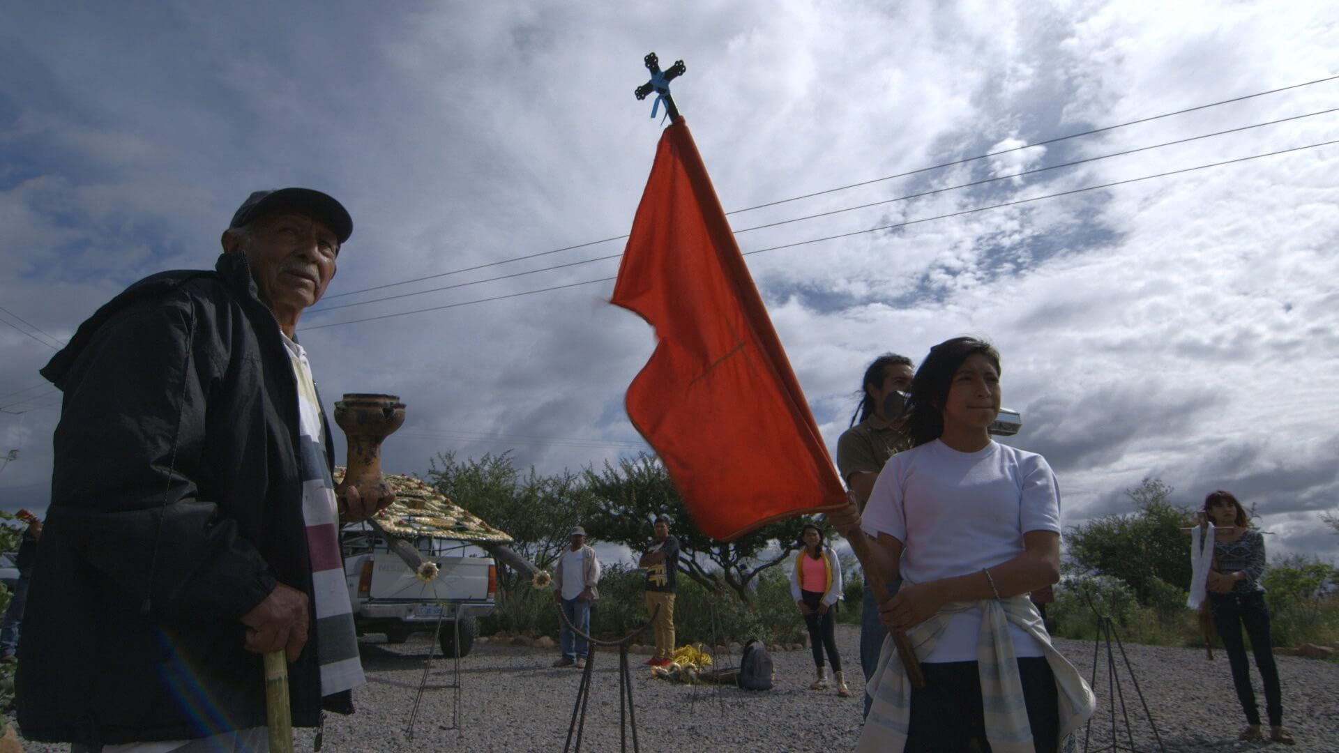 A still from The Age of Water. A person in a white T-shirt is holding a tall red flag with a cross atop the mast. Four other people stand behind them, in a patch of gravel.