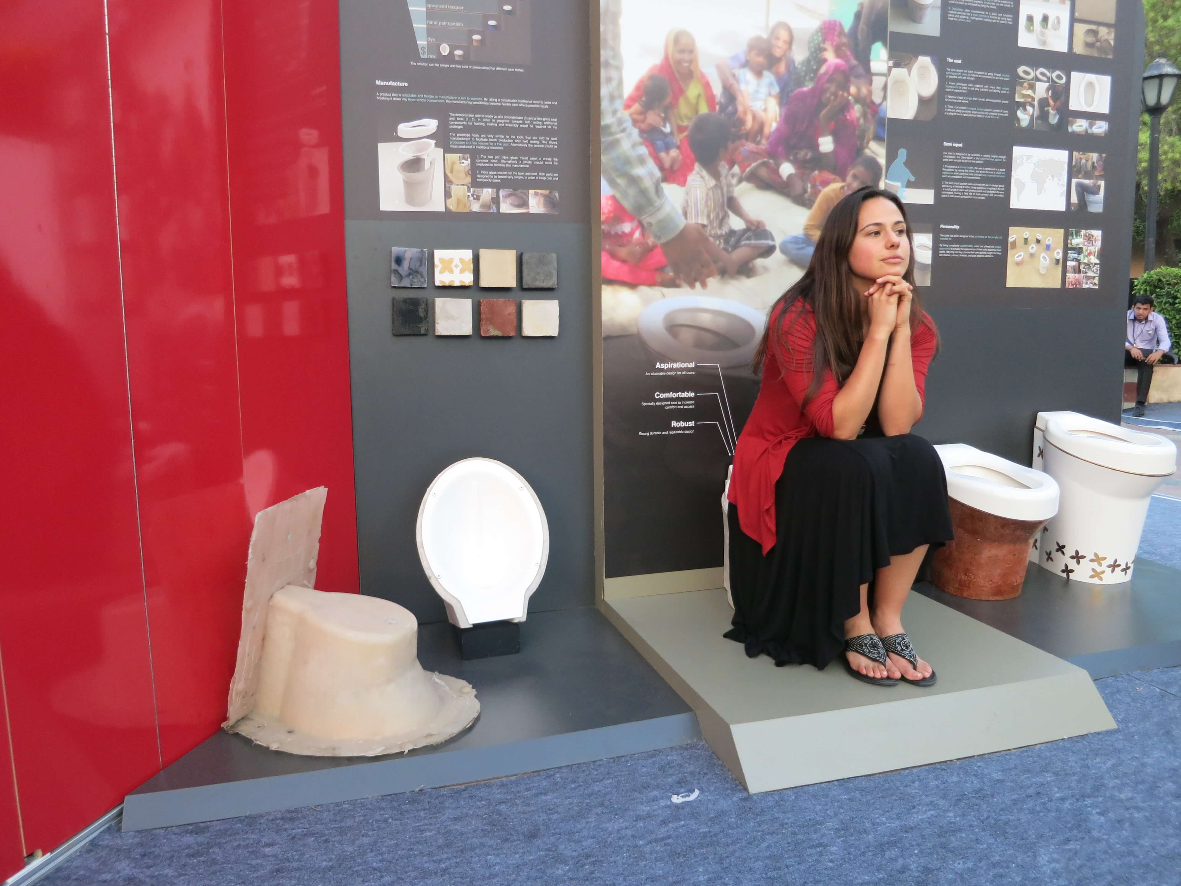 Still from Mister Toilet: The World's #2 Man. A woman sits on one of four toilets on display within an outdoor gallery. She is fully clothed with her elbows on her thighs and hands under her chin.