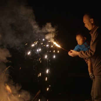 A still from The Departure. A man holds a small child in one arm, in the other hand, he holds a lit sparkler. It is nighttime.