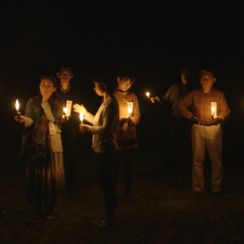 A still from The Departure. Six people hold candles, standing on a grass field in the middle of the night.