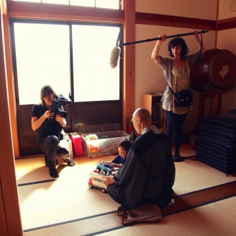 A still from The Departure. One person holds a camera and one person holds a boom. Both are pointed at a man and a child seated on the floor playing with a toy truck.