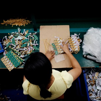Still from JOLIN: The Evolution of my Life. A woman is seen overhead as she disassembles electronic parts. Piles of the different parts surround her on her workspace table, in bins, and on the floor.