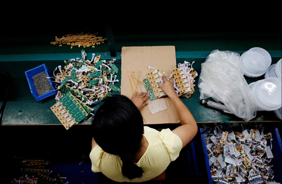 Still from JOLIN: The Evolution of my Life. A woman is seen overhead as she disassembles electronic parts. Piles of the different parts surround her on her workspace table, in bins, and on the floor.