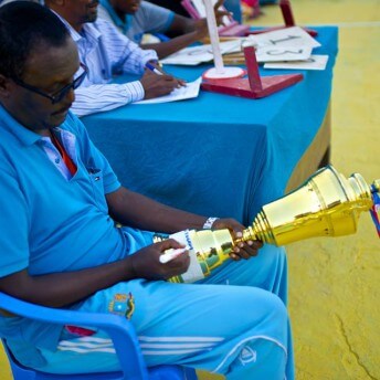 Still from Rajada Dalka/Nation's Hope. A man wearing a blue shirt and blue pants sits in a blue chair next to a table of two other men. He is writing on the bottom of a trophy.