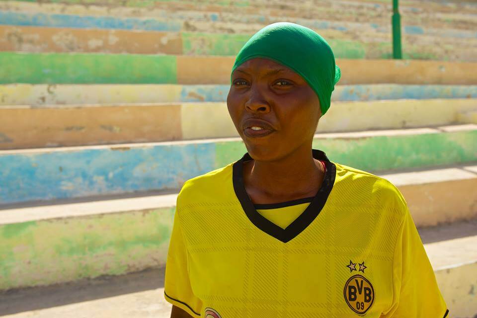 Still from Rajada Dalka/Nation's Hope. A woman stands in an exterior, in front of colorful, cement stadium-style seating steps. She is wearing a yellow jersey and a green headwrap.