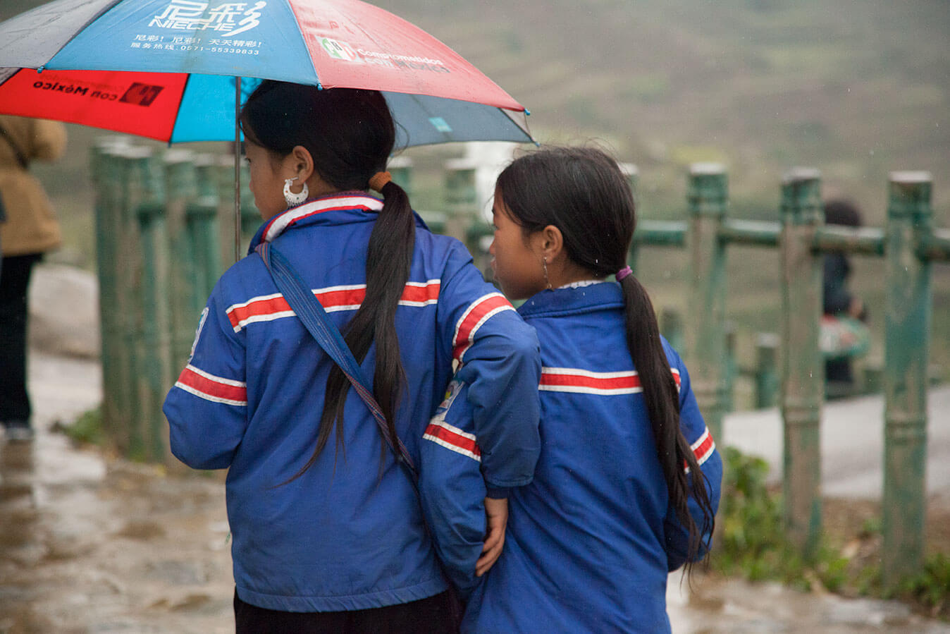 Still from Along the Line. Two young girls walk in the rain, one of them holds an umbrella, and both of them wear blue jackets.
