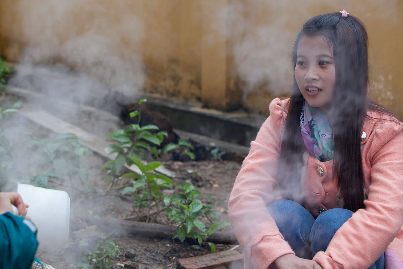 Still from Along the Line. A young girl is sitting on the ground, she is holding her knees with her arms, and is wearing a pink jacket and denim jeans. She is surrounded by smoke.