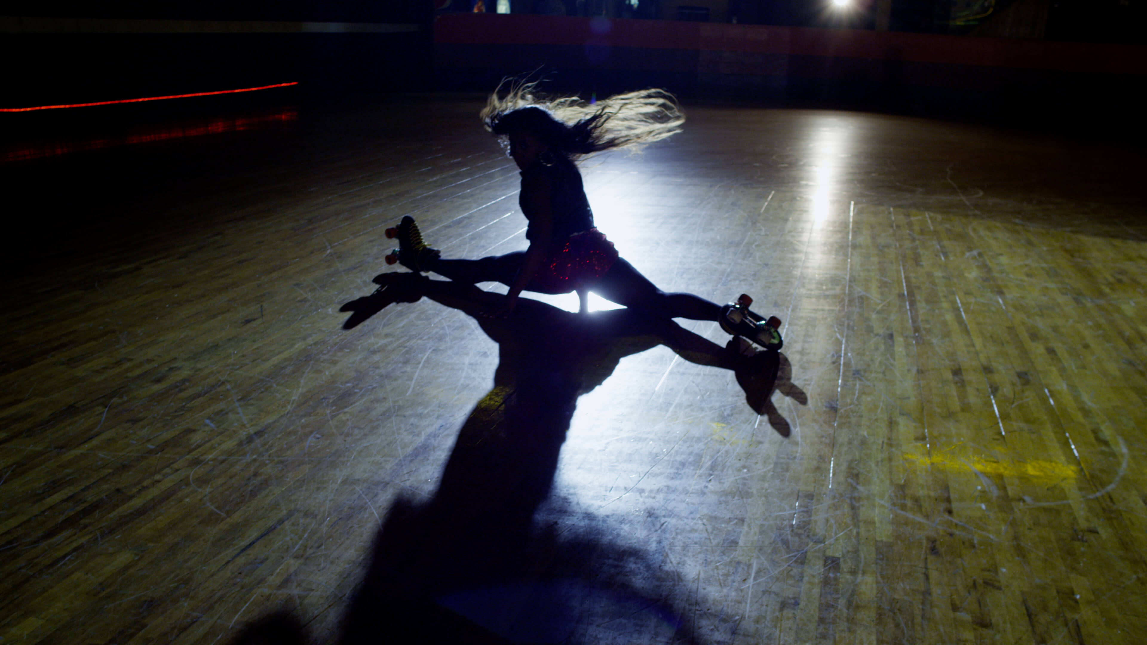 Still from United Skates. A woman in skates on a skating rink is doing the splits. The image is backlit.