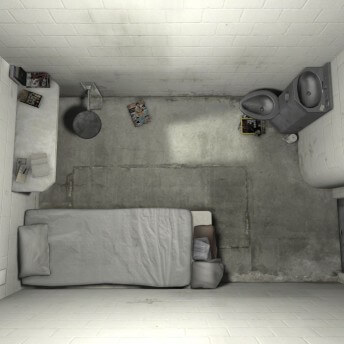 Still from An Immersive Experience of Solitary Confinement Impact & Innovation Initiative. Aerial shot of a prison cell: a bed with a bench, a shelf with some books, and a toilet, some magazines are spread over the floor.