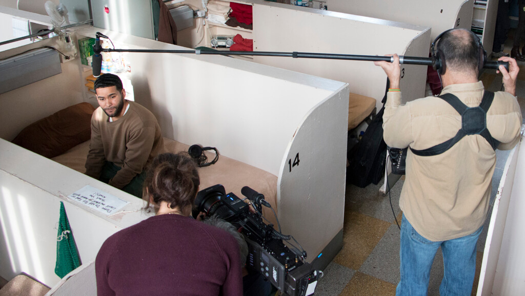 Production still of Lynn Novick's College Behind Bars: The Bard Prison Initiative. High angle shot of a woman behind the camera and the arm of a crew person holding a pole. A man is sitting on a white bench.