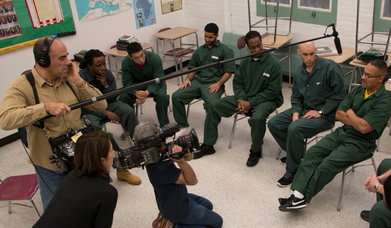 Production still of Lynn Novick's College Behind Bars: The Bard Prison Initiative. High angle shot of six men in prison uniforms sitting in a semi-circle inside of a classroom. A cameraman has a camera on his shoulder and films the men. Lynn the director is in the left corner looking at the shot. The audio pole is visible across the image.