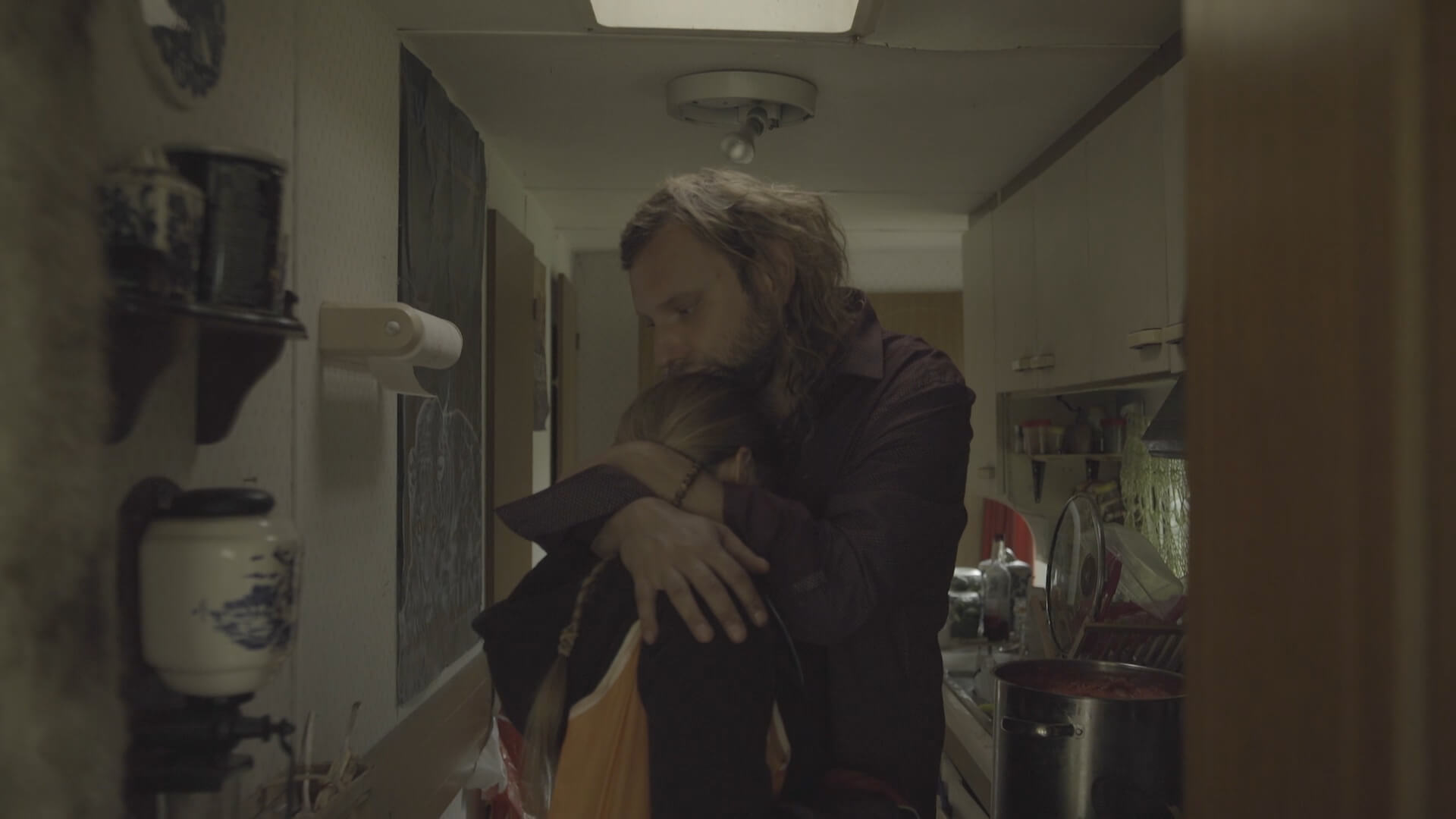 A blonde man and woman hugging in a small kitchen, with the woman's face hidden in the nook of the man's shoulder.