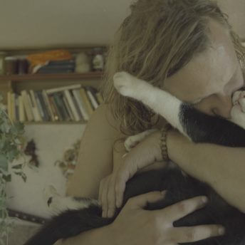 Close-up of a blonde person with long hair looking down, holding and kissing a black and white cat.