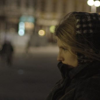 The profile of a woman from the shoulders up facing left, wearing a winter coat covering her mouth and head, standing in the street at night in front of blurry lights and buildings.