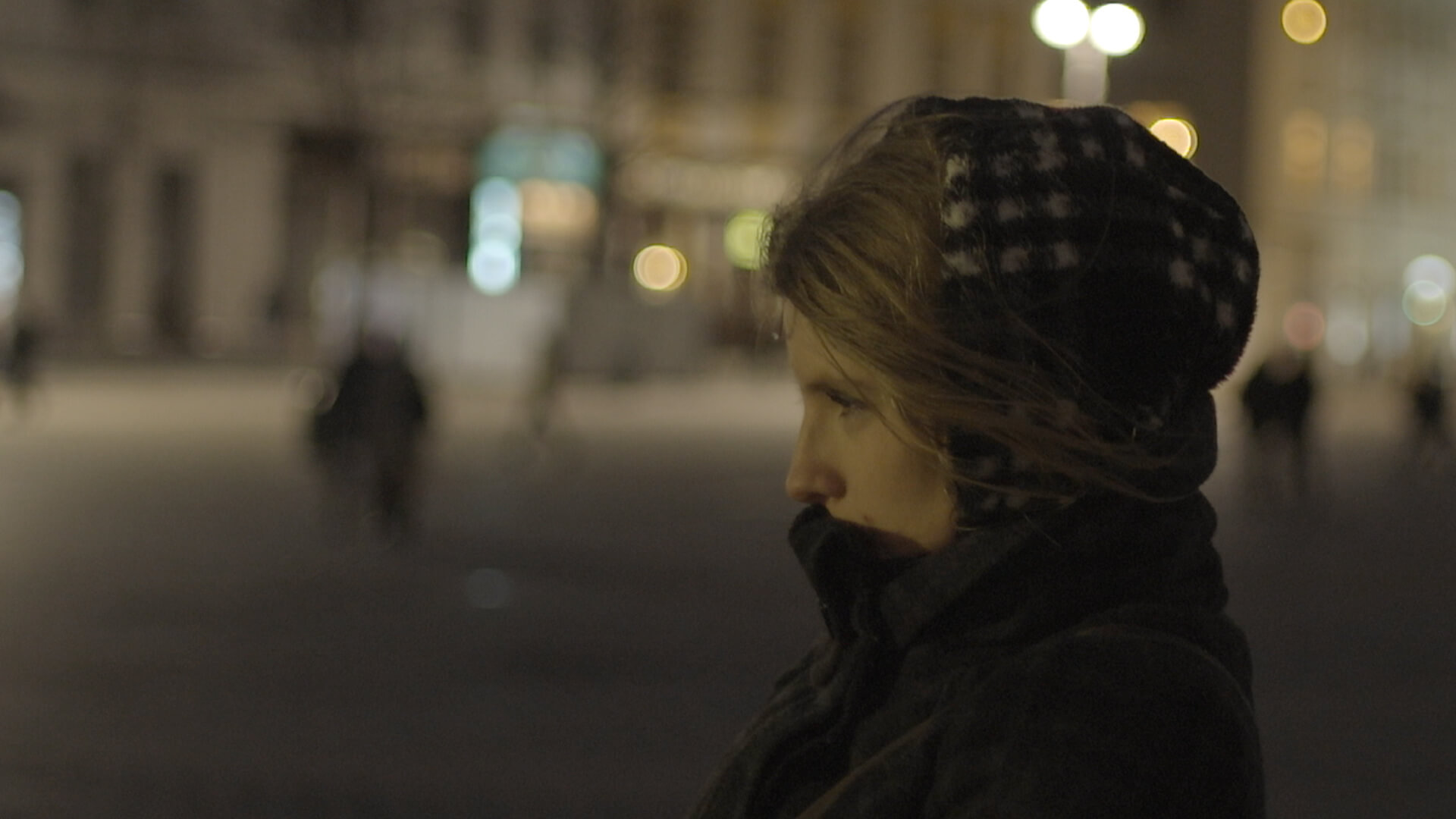 The profile of a woman from the shoulders up facing left, wearing a winter coat covering her mouth and head, standing in the street at night in front of blurry lights and buildings.