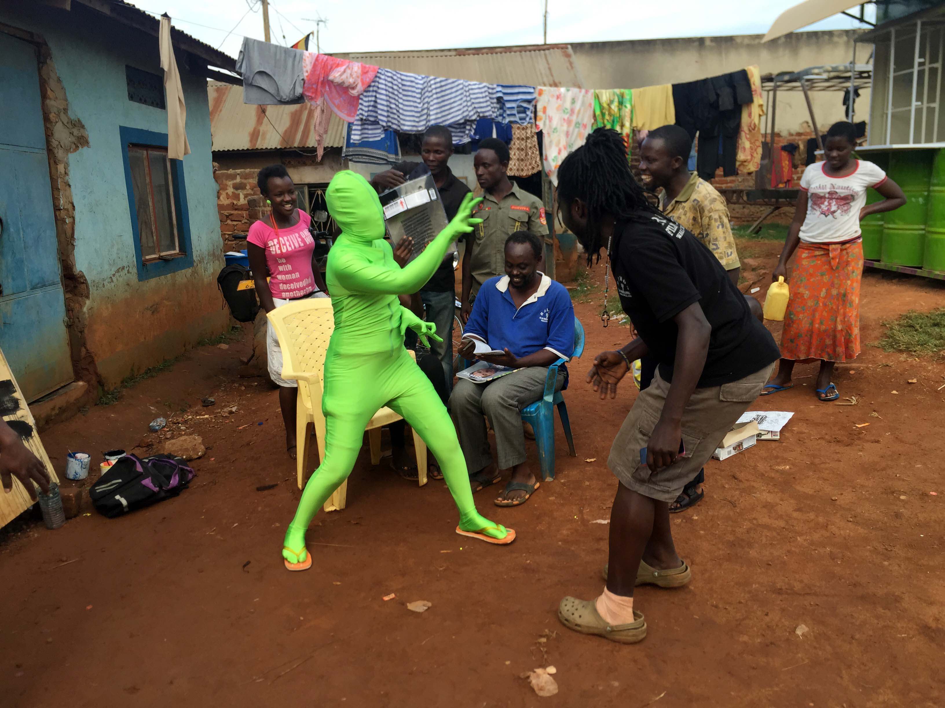 Still from Once Upon a Time in Uganda. A handful of people are gathered outside a couple houses. One person is sitting in a chair and another has a greenscreen suit. There is a clothing line full of clothes in the background.
