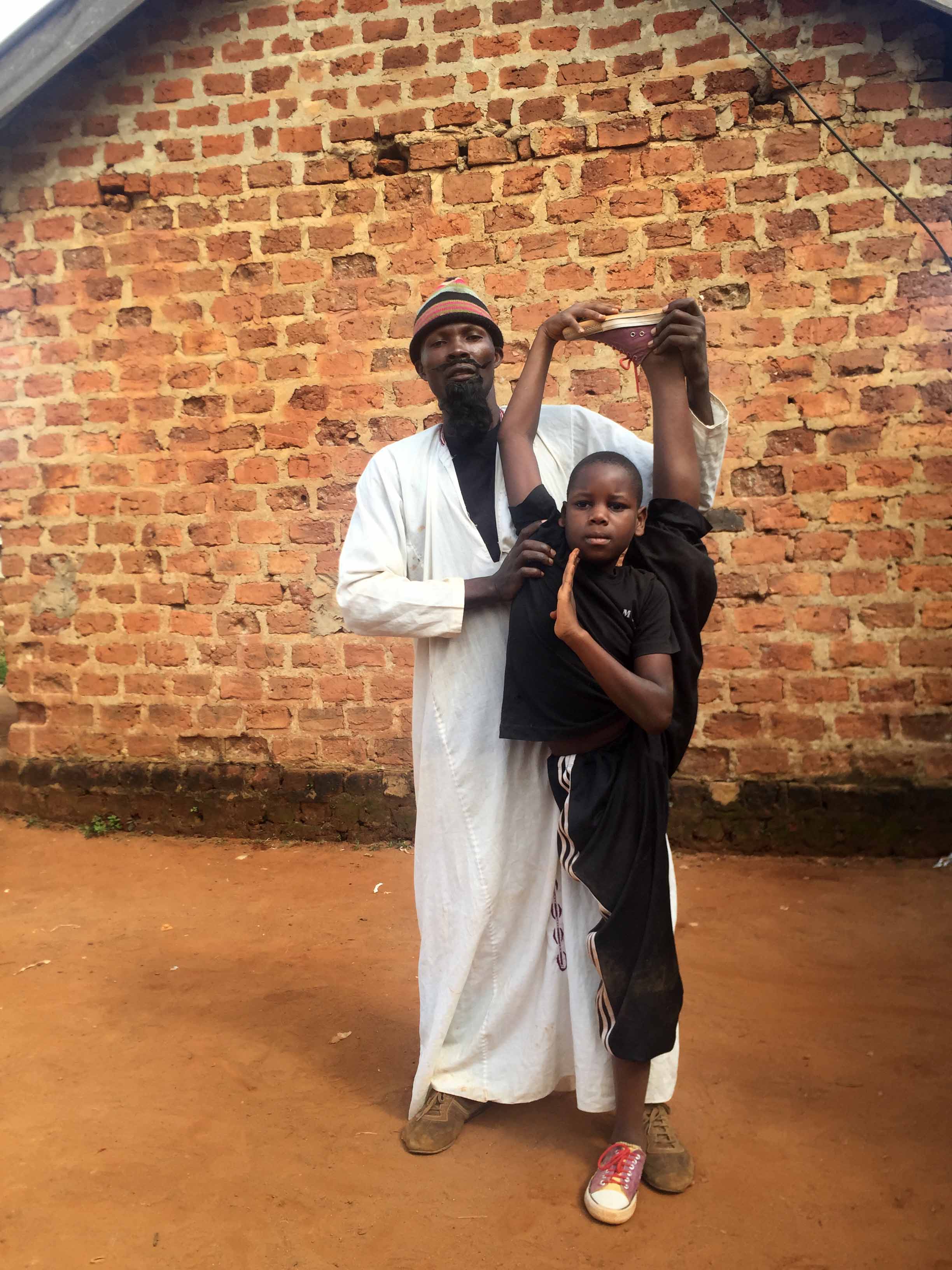 Still from Once Upon a Time in Uganda. A man stands behind and assists a young boy who is standing in a straddle position, standing on one foot with the other foot above his head. Behind them is a brick structure.